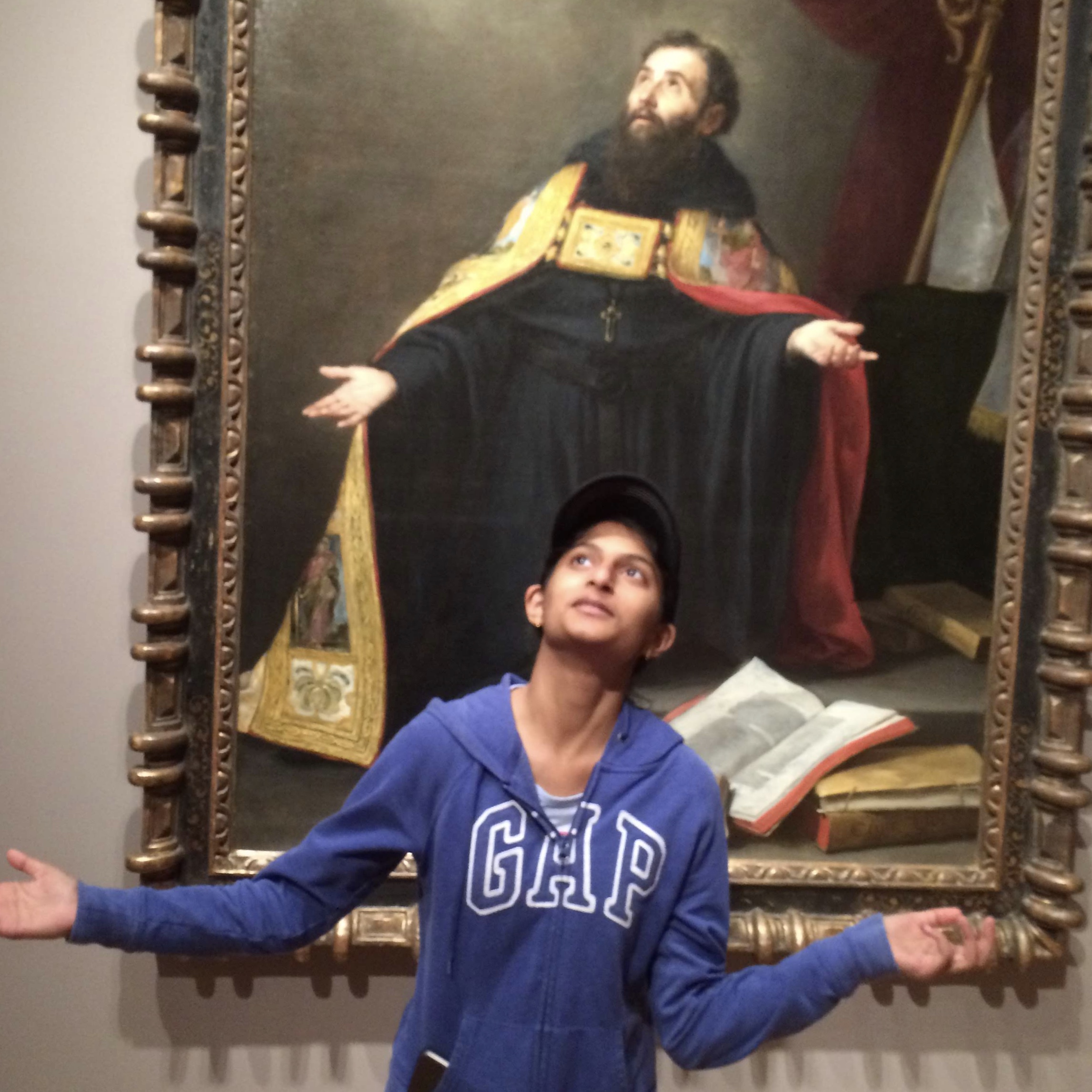 A medium brown skinned woman wearing a royal blue hoodie and a black hat with her arms out and looking upward in a questionable manner, mimicking a pose in the art piece directly behind her.